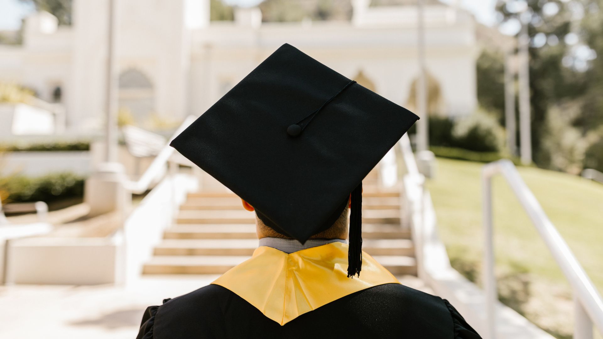 graduate in cap and gown walking up to the front steps of a college or university
